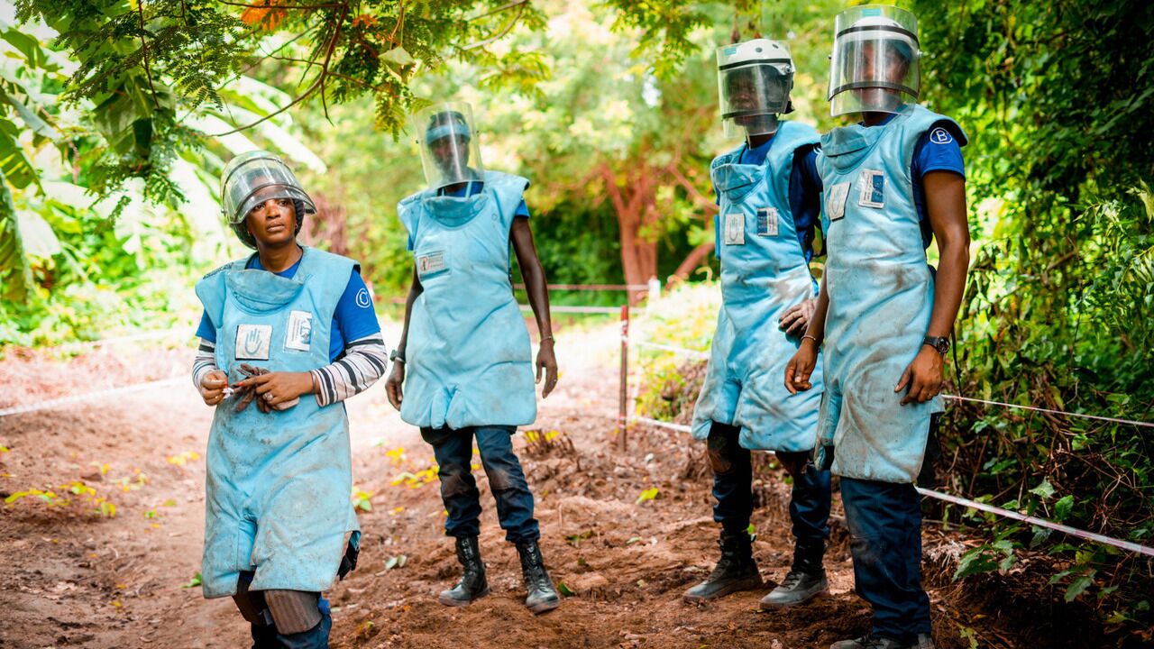 Four people dressed in protective mine clearance gear stand on a shady dirt track.