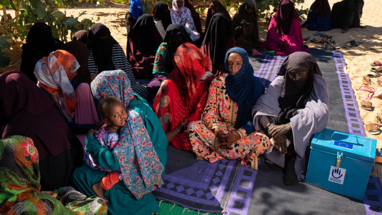 Several women are sitting on mats on the sand, waiting in the shade of the trees.